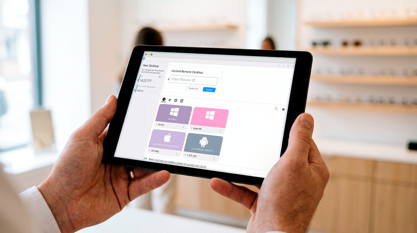 Hænder holder en tablet der viser et fjernsupport-værktøj med liste over forbundne enheder, i en moderne optikerbutik